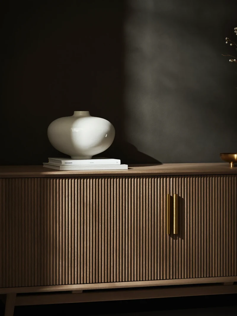Eichen-Sideboard mit Lamellenfront, Messinggriffen, Vase und Büchern vor dunkler Wand